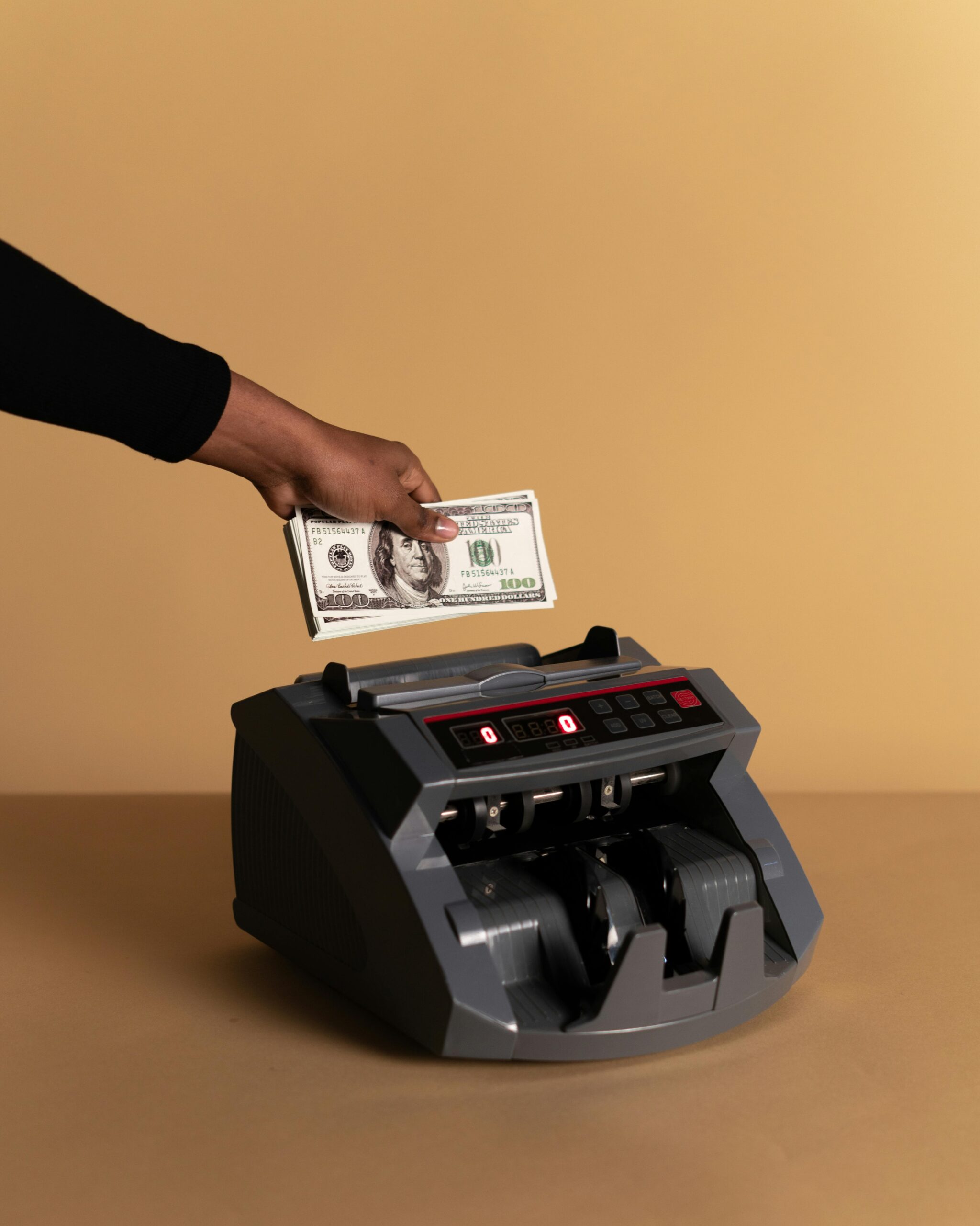 A hand holding us dollars in a studio setting with a money counting machine on a beige background.