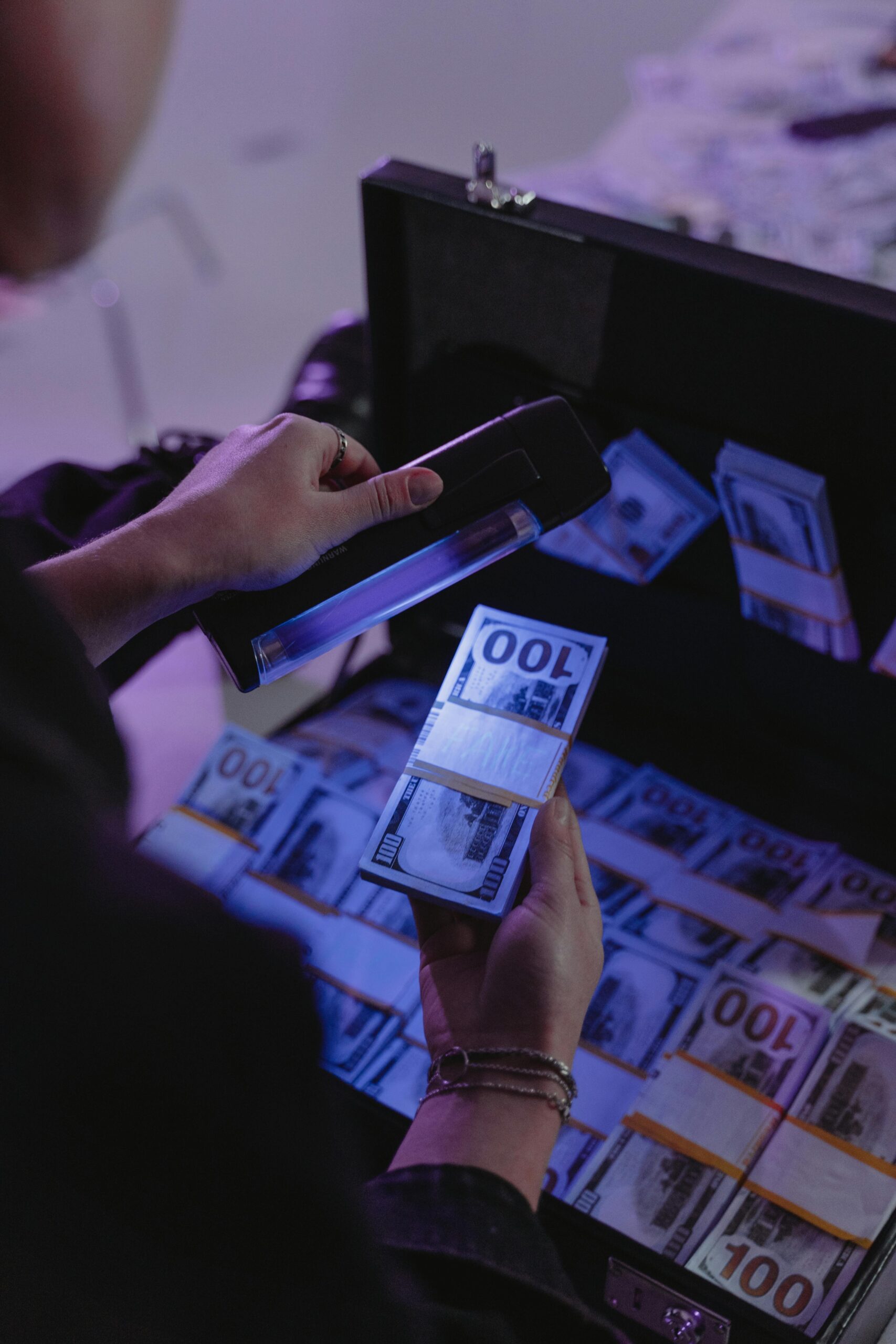 A person examining hundred dollar bills under UV light in a briefcase, ensuring authenticity.